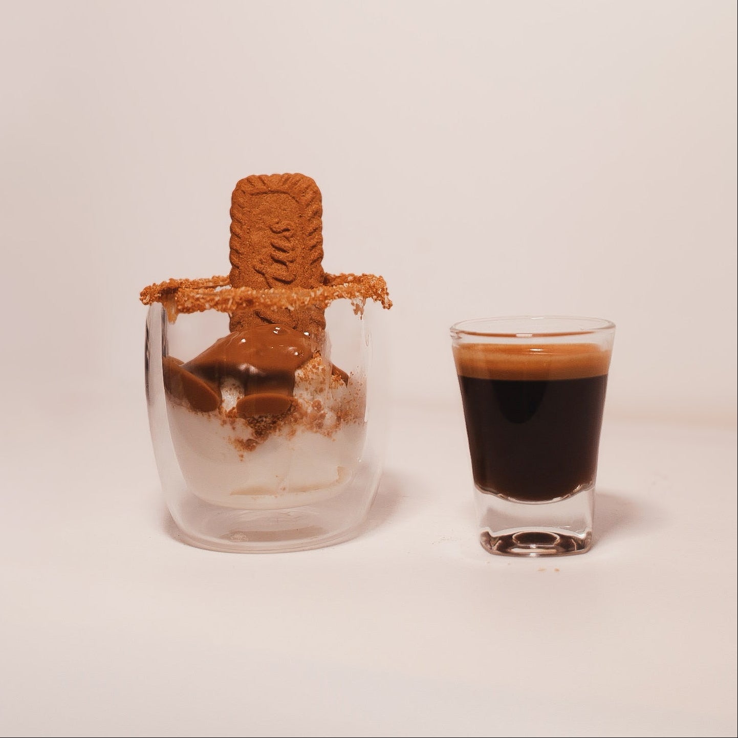 Small glass bottle with cork stopper next to a shot glass filled with dark liquid on a light pink background